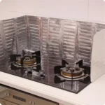 Silver folding aluminum oil splash guard placed around a frying pan on a gas stove to block grease splatter during cooking broccoli and other vegetables.