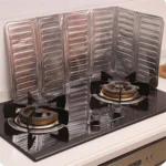Silver folding aluminum oil splash guard placed around a frying pan on a gas stove to block grease splatter during cooking broccoli and other vegetables.