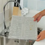 Silver folding aluminum oil splash guard placed around a frying pan on a gas stove to block grease splatter during cooking broccoli and other vegetables.