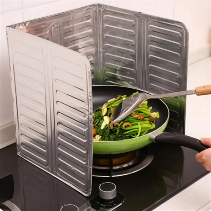 Silver folding aluminum oil splash guard placed around a frying pan on a gas stove to block grease splatter during cooking broccoli and other vegetables.